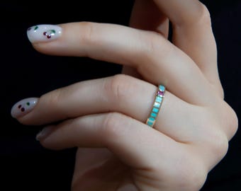 May include: A gold ring with a rectangular turquoise and ruby design. The ring is on a finger with white nail polish and small red and green dots. The background is black.