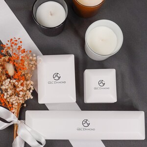 Puede incluir: Tres cajas blancas con el logotipo "GLC Diamond" en ellas. Las cajas est&aacute;n sobre una superficie gris con dos velas blancas y una vela marr&oacute;n.