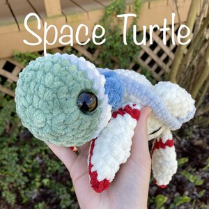 May include: A crocheted space turtle plush toy with a green shell, white and blue body, and red accents. The toy is being held in a hand and the text "Space Turtle PDF Pattern" is written below the toy.