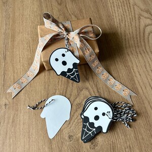 Op de afbeelding: Een ingepakt bruin cadeau met een lint met pompoenpatroon en een spookvormig label. Het label is wit met zwarte details, waaronder een gezicht en een spinnenwebontwerp. Extra labels en een bundel touw zijn ook zichtbaar.