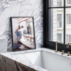 Pode incluir: Uma fotografia emoldurada de uma pessoa em uma casa de banho, repousando sobre uma bancada de m&aacute;rmore. A fotografia est&aacute; em uma moldura preta, e a casa de banho tem uma parede de m&aacute;rmore e uma janela com moldura preta. Ferragens douradas s&atilde;o vis&iacute;veis.