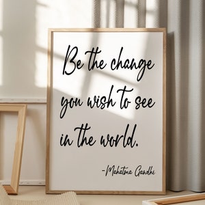 May include: A framed print with a white background and black text that reads "Be the change you wish to see in the world. -Mahatma Gandhi". The print is sitting on a light brown surface.