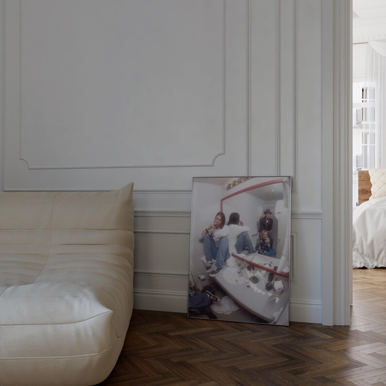 Pode incluir: Uma fotografia emoldurada, com uma moldura prateada, retratando pessoas em um banheiro, encostada em uma parede branca. A foto est&aacute; em um piso de madeira. Um sof&aacute; branco &eacute; vis&iacute;vel &agrave; esquerda.