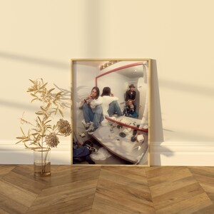 Pode incluir: Fotografia emoldurada com reflexo num espelho de pessoas numa casa de banho. O espelho tem uma moldura vermelha. As pessoas usam roupas casuais. A fotografia &eacute; exibida num ch&atilde;o de madeira junto a um vaso com ramos decorativos.