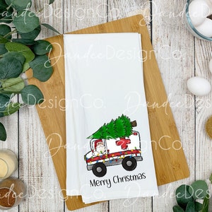 Pu&ograve; includere: Un asciugamano da cucina bianco con un'illustrazione di cartone animato di Babbo Natale che guida un'ambulanza con un albero di Natale in cima. L'ambulanza &egrave; decorata con luci di Natale. Il testo "Buon Natale" &egrave; stampato sotto l'immagine.
