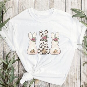 May include: White t-shirt with a graphic of three Easter bunnies. The middle bunny is wearing a leopard print pattern, while the bunnies on either side are white with pink and brown details. All three bunnies have pink flowers on their heads.