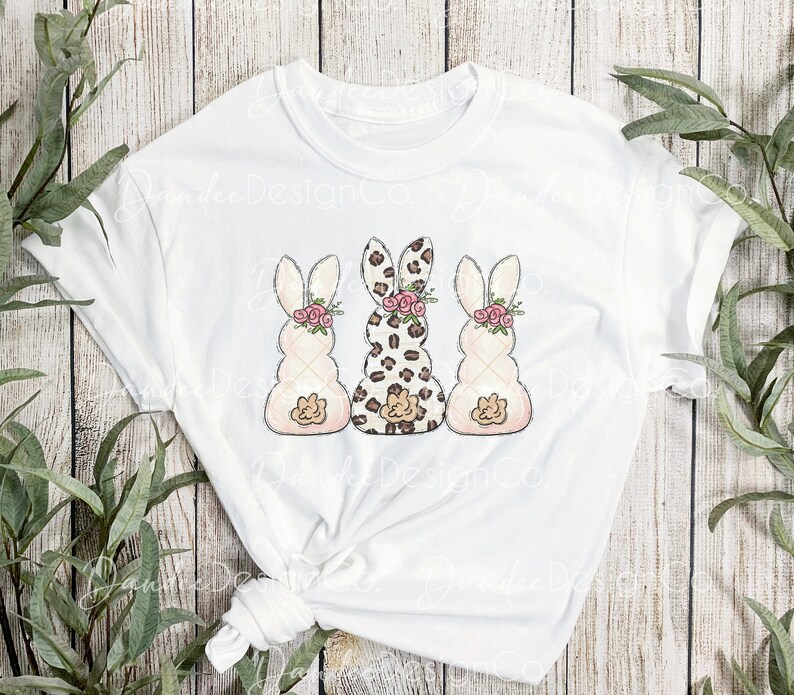 May include: White t-shirt with a graphic of three Easter bunnies. The middle bunny is wearing a leopard print pattern, while the bunnies on either side are white with pink and brown details. All three bunnies have pink flowers on their heads.