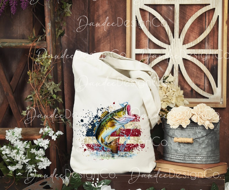May include: A white tote bag featuring a design of a largemouth bass leaping from the water against a backdrop of an American flag. The design is in shades of red, white, and blue.