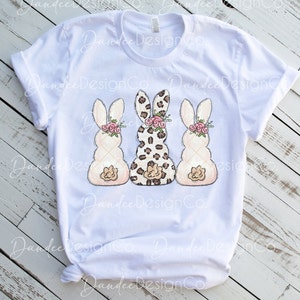 May include: Three Easter bunny-shaped figures, one with a leopard print pattern, and two with a white quilted pattern. Each bunny has a pink flower crown and a pink bow.