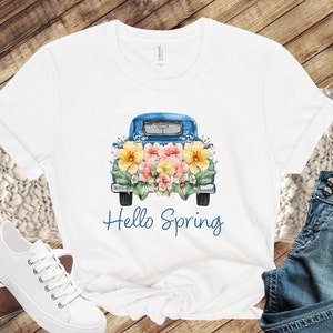 May include: White t-shirt with a blue pickup truck carrying a bouquet of yellow, pink, and white flowers. The text "Hello Spring" is printed below the truck.