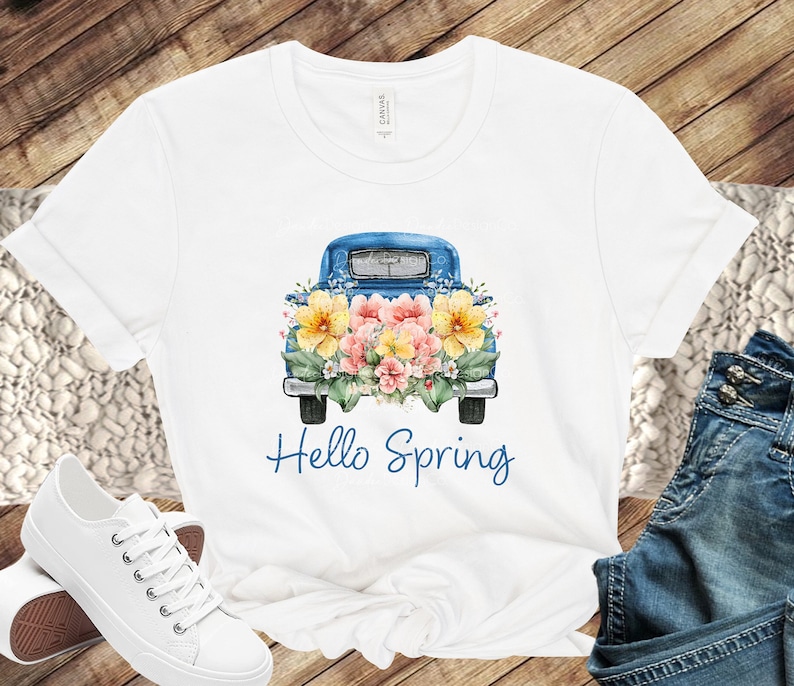 May include: White t-shirt with a blue pickup truck carrying a bouquet of yellow, pink, and white flowers. The text "Hello Spring" is printed below the truck.