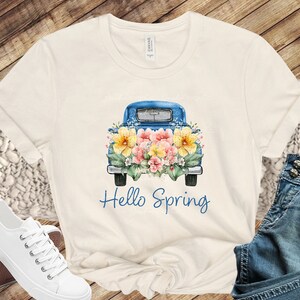 May include: A white t-shirt with a blue pickup truck carrying a bouquet of yellow and pink flowers. The text "Hello Spring" is printed below the truck.