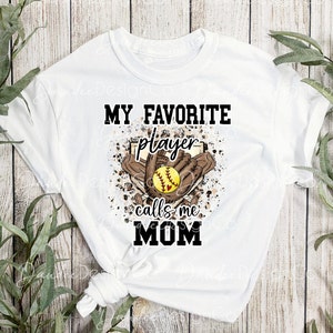 May include: White t-shirt with a brown and white graphic design. The graphic features a softball, a glove, and the text "My Favorite Player Calls Me Mom".