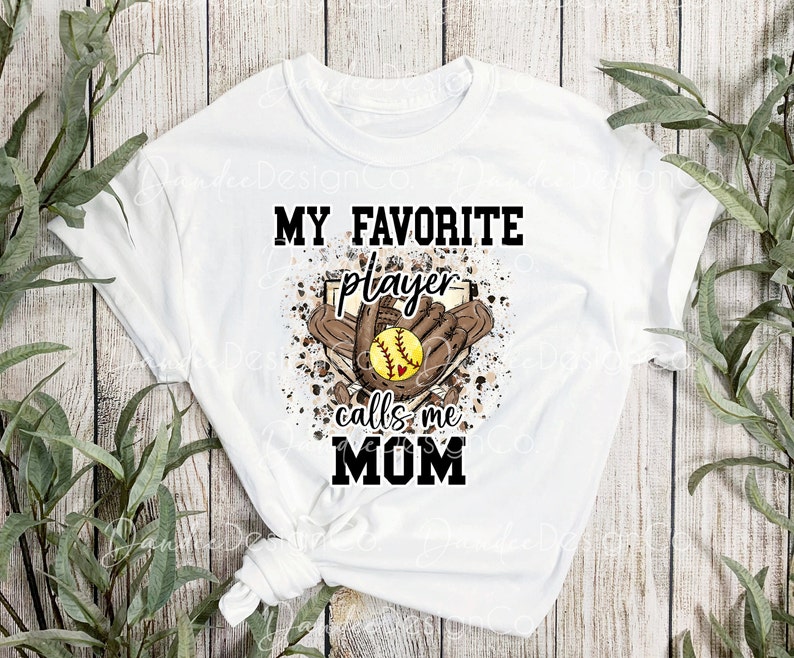 May include: White t-shirt with a brown and white graphic design. The graphic features a softball, a glove, and the text "My Favorite Player Calls Me Mom".