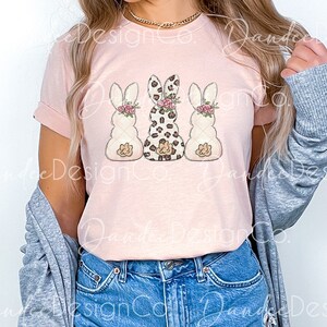 May include: A pink t-shirt with a graphic of three bunnies. The middle bunny is wearing a leopard print pattern and the other two bunnies are white with pink flowers.