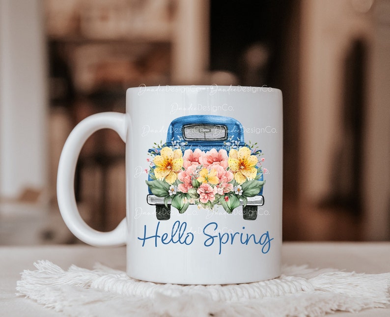 May include: White ceramic mug with a blue pickup truck filled with yellow and pink flowers. The text "Hello Spring" is printed on the mug.