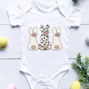 May include: White baby bodysuit with three bunny illustrations. The bunnies are in a row, with the middle bunny wearing a leopard print pattern and the bunnies on either side wearing a white and pink pattern. Each bunny has a pink flower headband.
