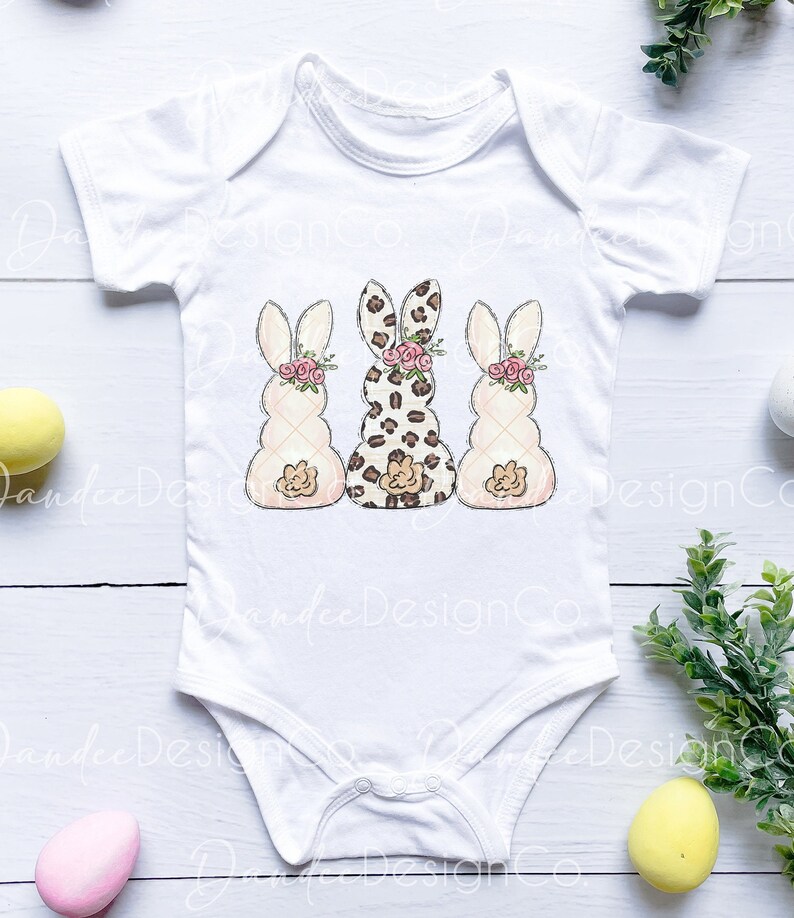 May include: White baby bodysuit with three bunny illustrations. The bunnies are in a row, with the middle bunny wearing a leopard print pattern and the bunnies on either side wearing a white and pink pattern. Each bunny has a pink flower headband.