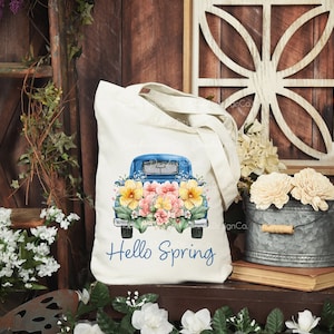 May include: A white tote bag with a blue pickup truck filled with colorful flowers. The truck has the text "Hello Spring" printed on the back.