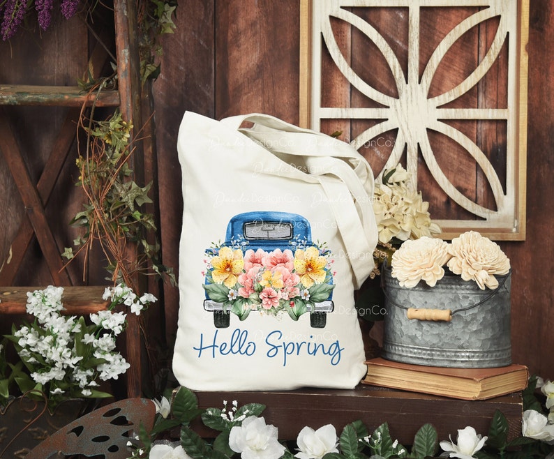 May include: A white tote bag with a blue pickup truck filled with colorful flowers. The truck has the text "Hello Spring" printed on the back.