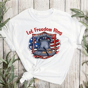 Puede incluir: Camiseta blanca con un dise&ntilde;o de bandera estadounidense roja, blanca y azul y una campana. El texto "Let Freedom Ring" est&aacute; escrito en rojo sobre la campana.