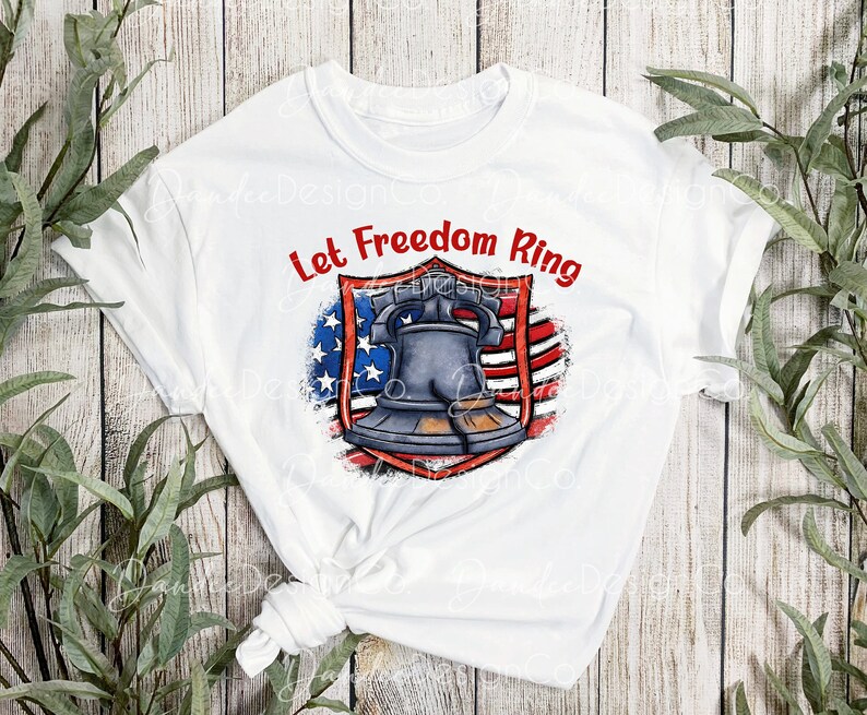 Puede incluir: Camiseta blanca con un dise&ntilde;o de bandera estadounidense roja, blanca y azul y una campana. El texto "Let Freedom Ring" est&aacute; escrito en rojo sobre la campana.