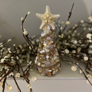 May include: A decorative seashell tree with a starfish topper. The tree is made of small, iridescent shell pieces and wrapped with twine. The tree is approximately 6 inches tall. White berry branches are in the background.