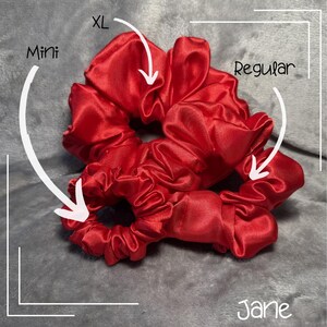 Puede incluir: Tres scrunchies de satén rojo en diferentes tamaños: mini, regular y XL. Los scrunchies están dispuestos en una pila, con el scrunchie mini en la parte inferior, el scrunchie regular en el medio y el scrunchie XL en la parte superior. Los scrunchies están hechos del mismo tejido de satén rojo y tienen una sensación suave y lujosa.