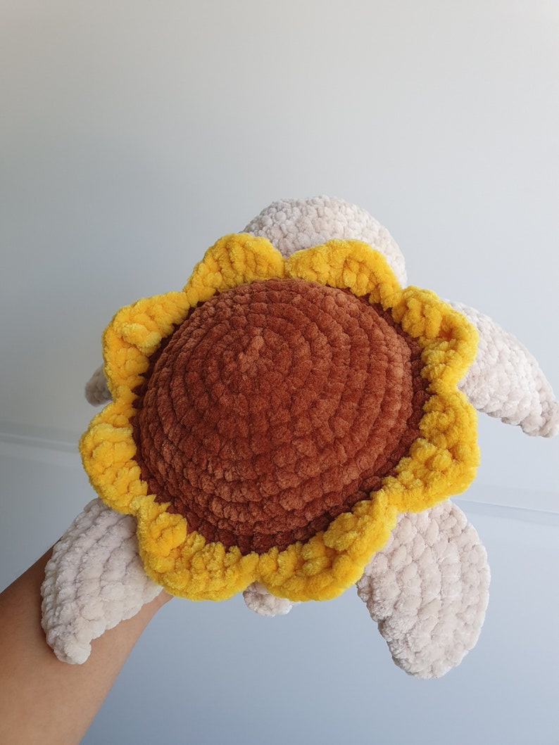 May include: A crocheted sunflower-shaped plush toy with a brown center and yellow petals. The toy has two beige wings.
