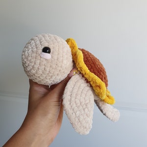 May include: A crocheted plush toy turtle with a brown and yellow shell. The turtle has a beige body and black eyes.