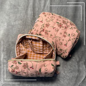 Puede incluir: Dos neceseres acolchados con estampado floral rosa. Una bolsa está abierta, revelando un interior de cuadros vichy con bolsillos. El exterior presenta un delicado estampado floral en tonos marrones y verdes. Las bolsas están diseñadas con cierre de cremallera y un asa pequeña.