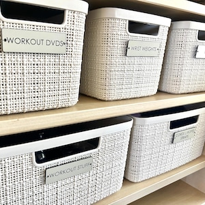 May include: White woven storage baskets with labels reading "WORKOUT DVDS", "WRIST WEIGHTS", "GRIPS", "WORKOUT STUFF", and "CORDS & ROPE STRAPS". The baskets are organized on wooden shelves.