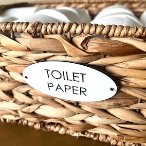 May include: A rectangular woven seagrass basket filled with white toilet paper rolls. An oval white metal plaque is attached to the front of the basket, featuring the black printed text TOILET PAPER in a clean, sans-serif font style.