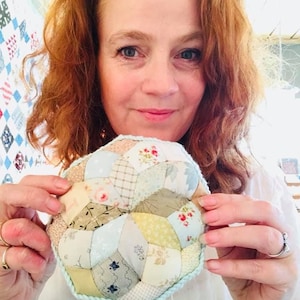 May include: A woman is holding a handmade pincushion with a blue and white crocheted border. The pincushion is made of patchwork fabric in shades of green, brown, and pink.