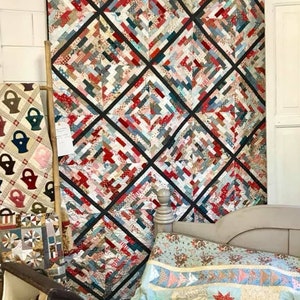 May include: A large patchwork quilt with a design featuring red, white and blue fabrics. The quilt is hanging on a wall in a room with a bed and other quilts.