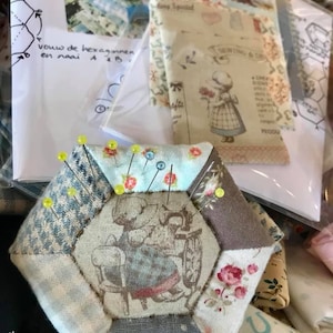 May include: A hexagonal pincushion made of patchwork fabric with a central panel featuring a girl sewing. The pincushion is filled with pins and is surrounded by other fabric scraps.