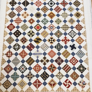 Op de afbeelding: Een wit quilt met een blauw ruitpatroon. Elke ruit bevat een ander kleurrijk patchwork-ontwerp.