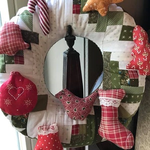 May include: A Christmas wreath made of patchwork fabric with various red, green and white ornaments, including a candy cane, a star, a bird, a stocking and a mitten.
