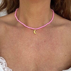Pu&ograve; includere: Una collana di perline rosa con un ciondolo a mezzaluna dorata. La collana &egrave; composta da piccole perline rosa chiaro. Il ciondolo dorato pende al centro della collana, indossata intorno al collo.