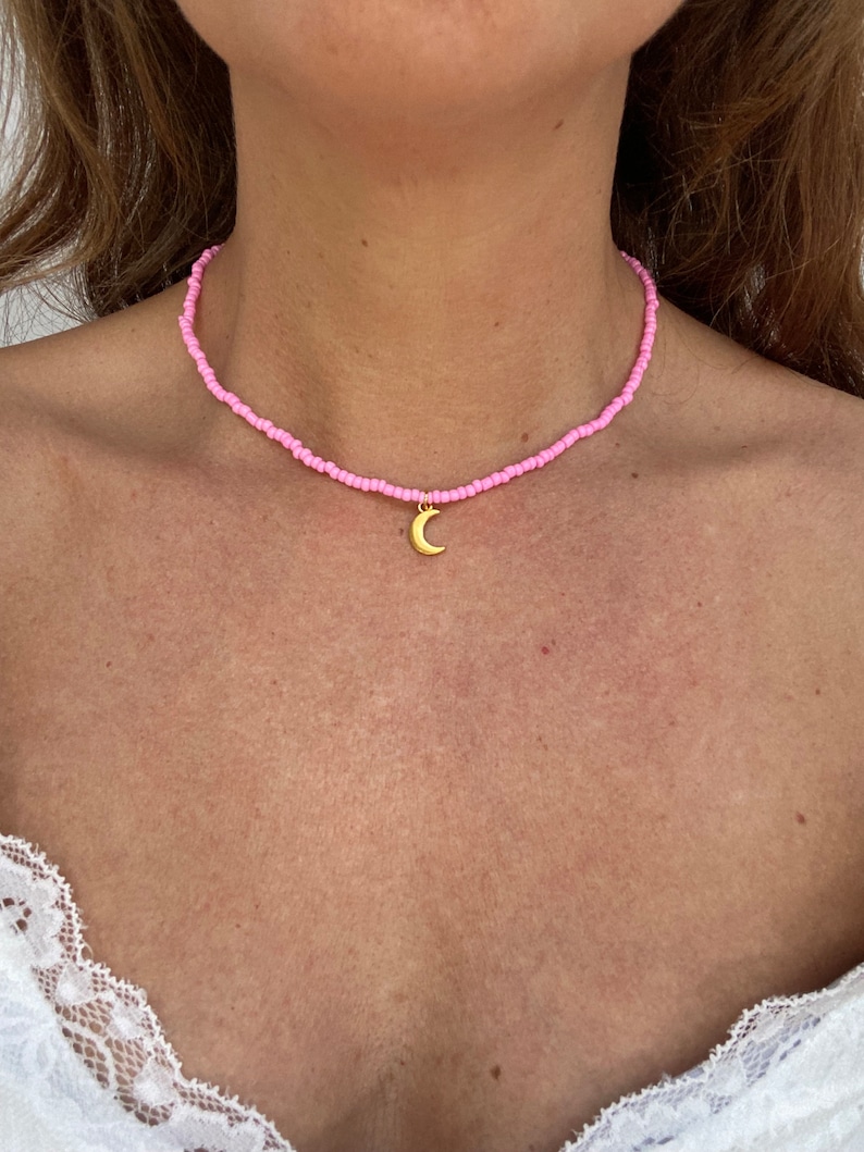 Pu&ograve; includere: Una collana di perline rosa con un ciondolo a mezzaluna dorata. La collana &egrave; composta da piccole perline rosa chiaro. Il ciondolo dorato pende al centro della collana, indossata intorno al collo.