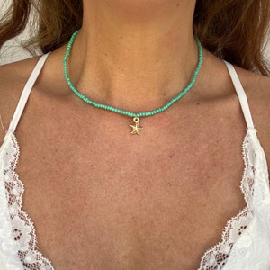 Pu&ograve; includere: Una collana girocollo con perline turchesi con un ciondolo a forma di stella marina color oro. La collana &egrave; indossata intorno al collo. Lo sfondo &egrave; un top bianco.