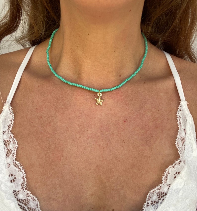 Pu&ograve; includere: Una collana girocollo con perline turchesi con un ciondolo a forma di stella marina color oro. La collana &egrave; indossata intorno al collo. Lo sfondo &egrave; un top bianco.