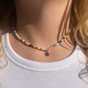 Pu&ograve; includere: Una collana di perline colorate con un piccolo ciondolo a forma di stella argentata. La collana presenta piccole perline rotonde nei toni del rosa, giallo, arancione, blu e viola. La collana &egrave; indossata sopra una canotta bianca a coste.