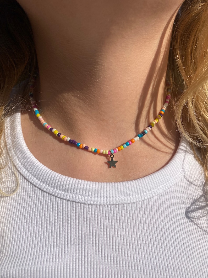 Pu&ograve; includere: Una collana di perline colorate con un piccolo ciondolo a forma di stella argentata. La collana presenta piccole perline rotonde nei toni del rosa, giallo, arancione, blu e viola. La collana &egrave; indossata sopra una canotta bianca a coste.