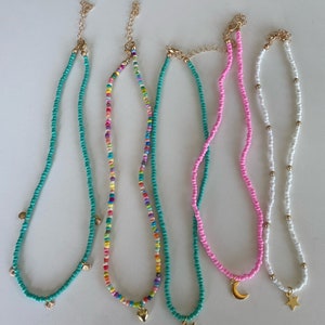 Pu&ograve; includere: Una collezione di collane di perline in vari colori. Le collane presentano ciondoli color oro, tra cui conchiglie, un cuore, una stella e una luna crescente. Le perline sono turchesi, rosa, arcobaleno e bianche.
