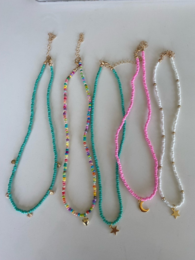 Pu&ograve; includere: Una collezione di collane di perline in vari colori. Le collane presentano ciondoli color oro, tra cui conchiglie, un cuore, una stella e una luna crescente. Le perline sono turchesi, rosa, arcobaleno e bianche.