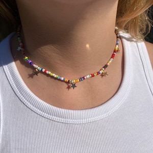 Pu&ograve; includere: Una collana di perline colorate con ciondoli a forma di stella dorata. La collana presenta piccole perline rotonde in vari colori, tra cui rosso, giallo, blu e bianco. &Egrave; indossata sopra una canotta bianca.