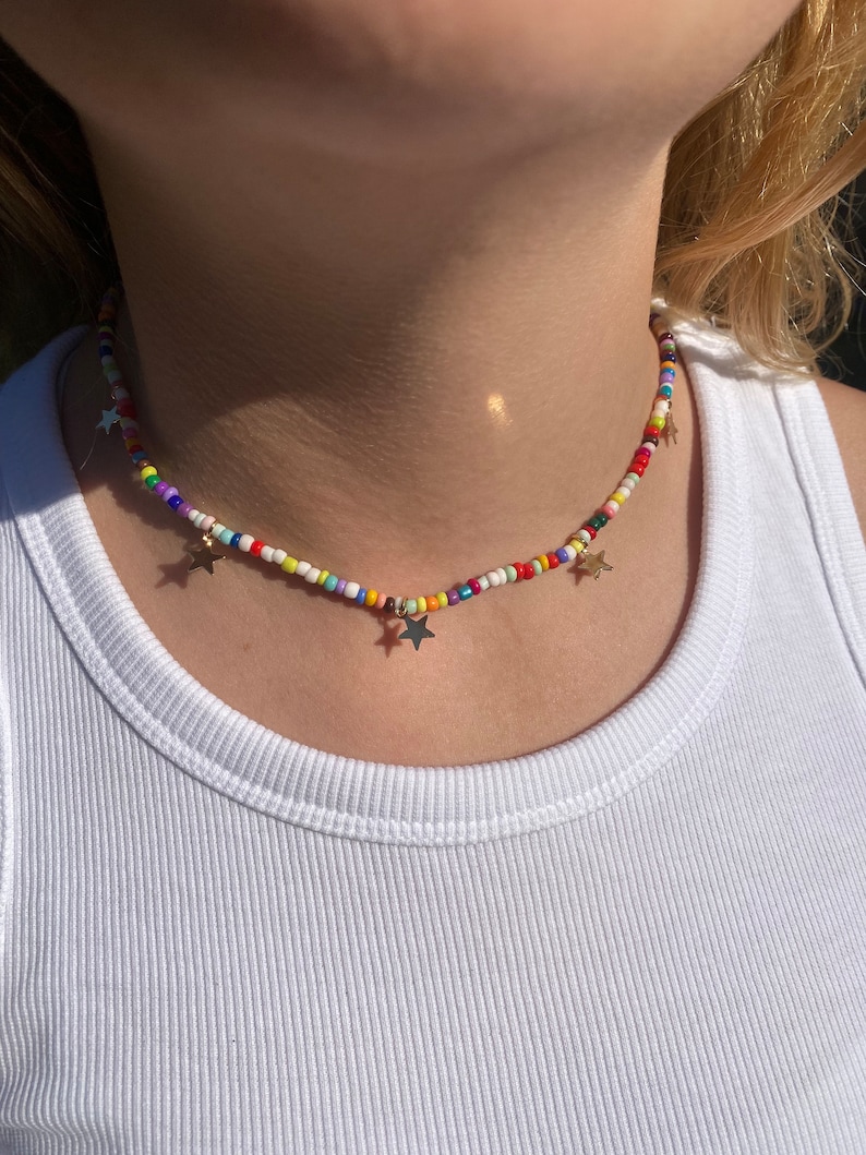Pu&ograve; includere: Una collana di perline colorate con ciondoli a forma di stella dorata. La collana presenta piccole perline rotonde in vari colori, tra cui rosso, giallo, blu e bianco. &Egrave; indossata sopra una canotta bianca.
