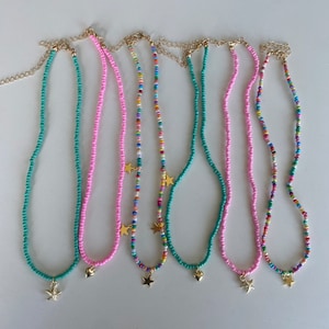 Pu&ograve; includere: Una collezione di collane di perline in vari colori, tra cui turchese, rosa e fili multicolore. Ogni collana presenta un ciondolo color oro, come una stella o un cuore, e una catena color oro.