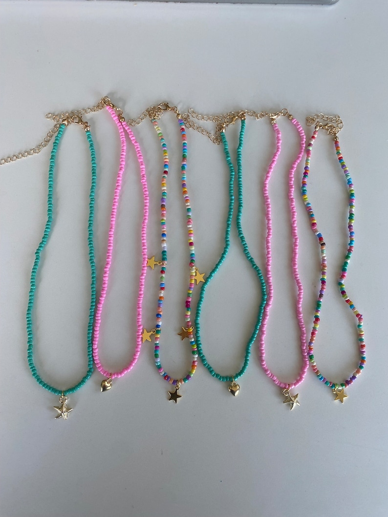 Pu&ograve; includere: Una collezione di collane di perline in vari colori, tra cui turchese, rosa e fili multicolore. Ogni collana presenta un ciondolo color oro, come una stella o un cuore, e una catena color oro.